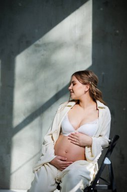 Beyaz takım elbiseli şık hamile bir kadın sandalyeye oturur ve nazikçe karnına dokunur. Doğumu bekliyorum. Bakım ve annelik. Sevgi ve şefkat.