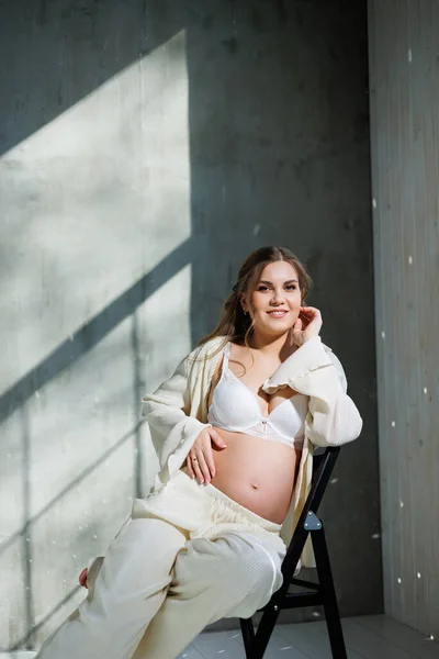 白いスーツを着たスタイリッシュな妊婦さんが椅子に座り 優しい笑顔でお腹に触れます 出産を待っている 気をつけてね 愛と優しさ — ストック写真