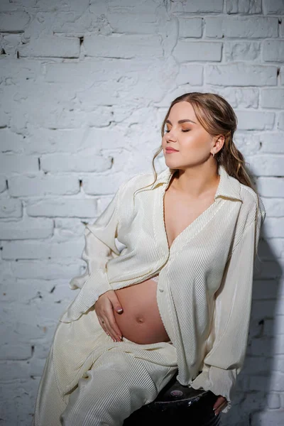 白いスーツの妊婦さんが白いレンガの壁に座り 優しい笑顔でお腹に触れています 出産を待っている 気をつけてね 愛と優しさ — ストック写真
