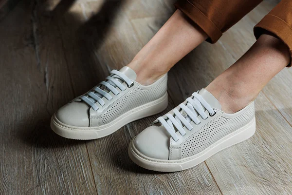 Stock image Close-up of female legs in leather white shoes. Young woman standing in leather stylish white sneakers in brown pants. Summer women's shoes. Casual street style.