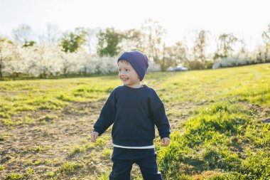 Küçük bir çocuk, 3 yaşında, kazak ve şapka giymiş, çiçek açan bir bahçede koşuyor. 3 yaşındaki çocuklar için giysiler. Mutlu bir Eotian çocuğu çiçek açan elma ağaçlarının arasında beyazdır.