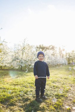Neşeli çocuk. 3 yaşında, çiçek açan bir bahçede yürüyüşe çıkmış. Küçük bir çocuk, 3 yaşında, kazak ve şapka giymiş, çiçek açan bir bahçede koşuyor. 3 yaşındaki çocuklar için giysiler. Çocuk sevincinin duyguları
