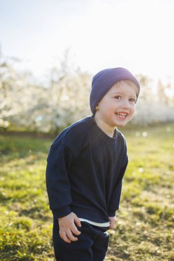 Neşeli çocuk. 3 yaşında, çiçek açan bir bahçede yürüyüşe çıkmış. Küçük bir çocuk, 3 yaşında, kazak ve şapka giymiş, çiçek açan bir bahçede koşuyor. 3 yaşındaki çocuklar için giysiler. Çocuk sevincinin duyguları