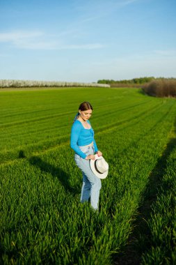 Kotlu ve şapkalı hoş bir kadın yeşil bir tarlada duruyor. Mavi bluzlu ve kot pantolonlu gülümseyen bir kadın yeşil çimenlerde yürür..