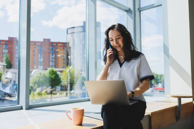 Diz üstü bilgisayarı, akıllı telefonu, kahve fincanı olan neşeli bir kadın, dizüstü bilgisayara bakıyor ve gündüzleri kafede otururken notlar alıyor. Uzaktan kumandayla çalışan bir kadın.