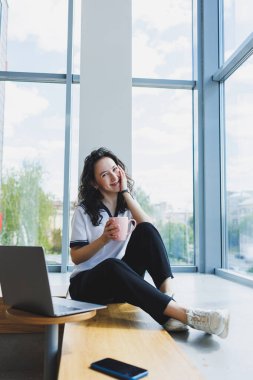 Gülümseyen kıvırcık saçlı kadın bir kafede oturuyor, bilgisayarın başında çalışıyor, akıllı telefondan mesajlar okuyor. Uzak çalışma