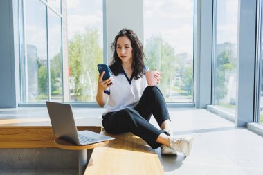 Gülümseyen kıvırcık saçlı kadın bir kafede oturuyor, bilgisayarın başında çalışıyor, akıllı telefondan mesajlar okuyor. Uzak çalışma