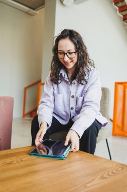 Başarılı bir kadın, günlük giysiler içinde gözlük takıyor modern kafedeki rahat koltukta oturuyor ve tablet internette çalışıyor. Uzaktan kumanda. Bağımsız bir çalışanın işi