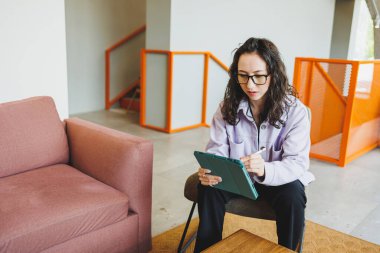 Başarılı bir kadın, günlük giysiler içinde gözlük takıyor modern kafedeki rahat koltukta oturuyor ve tablet internette çalışıyor. Uzaktan kumanda. Bağımsız bir çalışanın işi