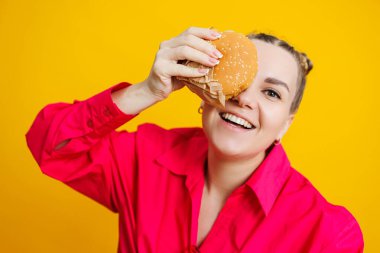 Hamburger yiyen komik hamile genç kadın. Sarı stüdyo duvarında izole bir şekilde dururken elinde lezzetli bir sandviç tutan hamile kadın. Abur cubur konsepti. Hamilelik süresince fast food.