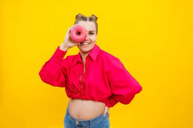 Sarı arka planda izole edilmiş pembe tişörtlü neşeli hamile bir kadın yüzünde neşeli bir ifadeyle donut tutuyor. Hamilelik sırasında tatlı yiyecekler.