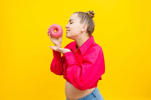 Smiling pregnant woman in pink shirt holding delicious sweet donuts on isolated yellow background. Sweet caloric food during pregnancy. A pregnant woman eats fatty pastries