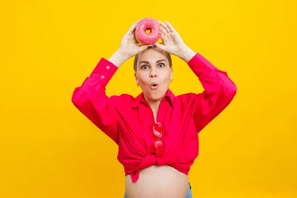 stock image Smiling pregnant woman in pink shirt holding delicious sweet donuts on isolated yellow background. Sweet caloric food during pregnancy. A pregnant woman eats fatty pastries