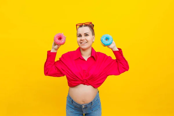 A smiling pregnant woman in a pink shirt holds donuts in her hands on a yellow background. Sweet food during pregnancy. Harmful food during pregnancy.