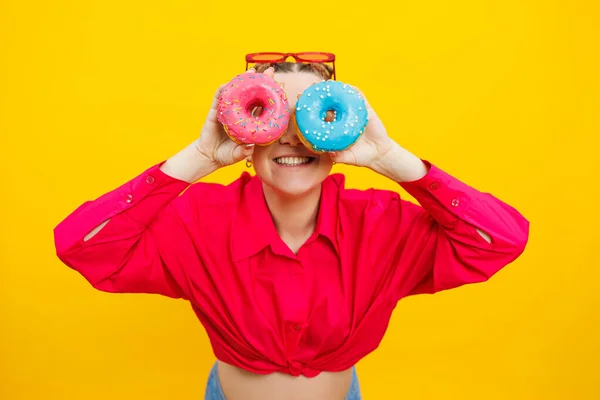 A smiling pregnant woman in a pink shirt holds donuts in her hands on a yellow background. Sweet food during pregnancy. Harmful food during pregnancy.