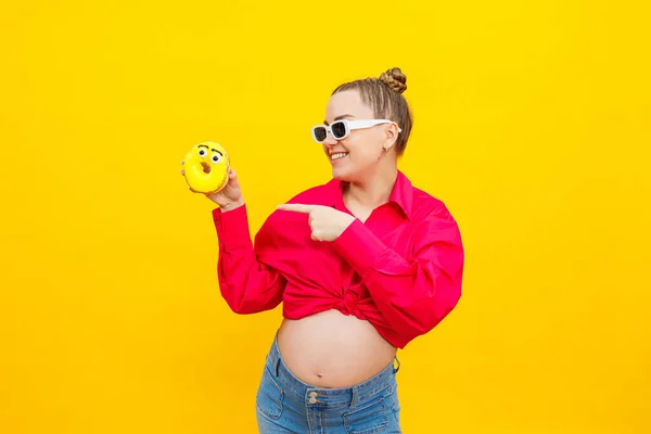 Positive pregnant woman eating donuts wearing pink shirt isolated on yellow background. Happiness from pregnancy while expecting a child. High quality photo. Harmful food during pregnancy