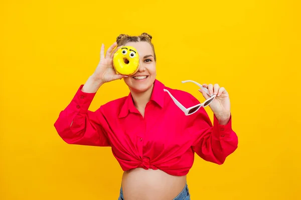 Positive pregnant woman eating donuts wearing pink shirt isolated on yellow background. Happiness from pregnancy while expecting a child. High quality photo. Harmful food during pregnancy