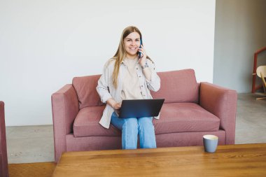 Modern kafeteryadaki rahat kanepede dizüstü bilgisayarlı ve telefonlu rahat koltukta oturan, cep telefonuyla konuşan serbest çalışan gülümseyen kadın. Uzak çalışma