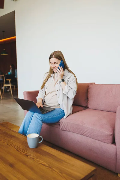 Smiling female freelancer in casual clothes sitting in comfortable sofa in modern cafeteria at table with laptop and phone, talking on mobile phone. Remote work