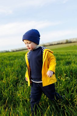 Sarı ceketli ve şapkalı üç yaşında şirin bir çocuk yeşil bir alanda koşuyor. Gülümseyen bir çocuk yeşil çimlerde koşar