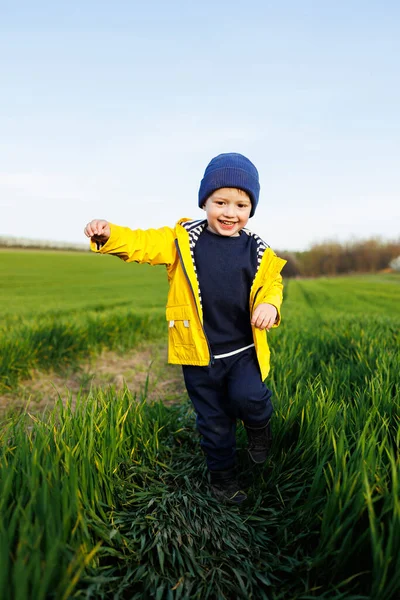 一个身穿黄色夹克头戴黄色帽子的可爱的三岁小男孩在绿地上奔跑 一个笑着的孩子在绿草上奔跑 免版税图库照片