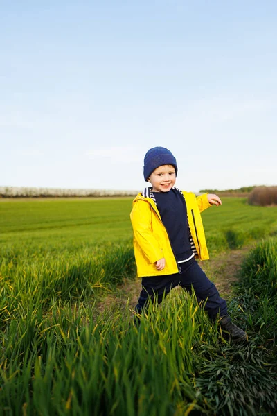 一个身穿黄色夹克头戴黄色帽子的可爱的三岁小男孩在绿地上奔跑 一个笑着的孩子在绿草上奔跑 — 图库照片