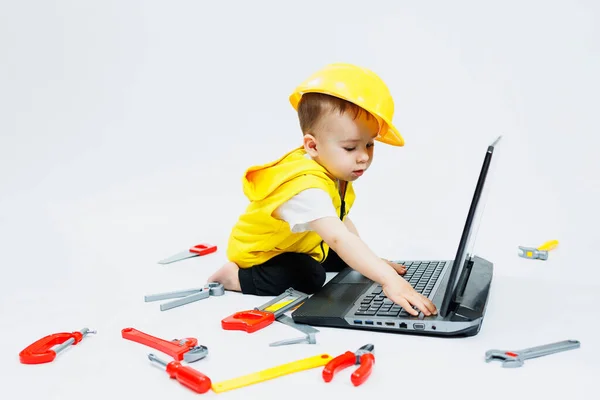 白い背景に黄色いベストを着た2歳の少年がラップトップに座ってプラスチック製の建設ツールで遊んでいます 小さな子供のためのおもちゃ ストック画像