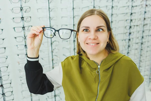 可愛い女性が眼鏡店で眼鏡を選ぶ 女性が眼鏡を買う 眼鏡の異なるモデルのお店の窓の背景に女性 — ストック写真