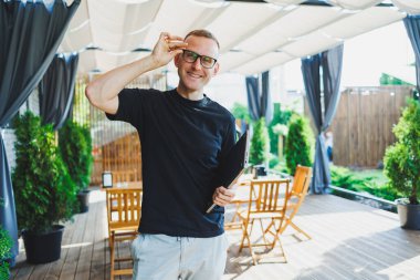 Gülümseyen genç bir adam elinde dizüstü bilgisayarla bir kafede bir terasta duruyor. Çevrimiçi çalış, serbest çalışan. Çevrimiçi uzak yaz işi.