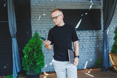 Şirin bir genç adam elinde dizüstü bilgisayarla bir kafede bir terasta duruyor ve telefonla konuşuyor. Çevrimiçi çalış, serbest çalışan. Çevrimiçi uzak yaz işi.