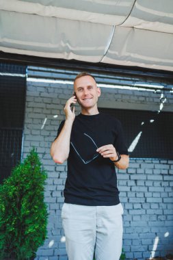 Şirin bir genç adam elinde dizüstü bilgisayarla bir kafede bir terasta duruyor ve telefonla konuşuyor. Çevrimiçi çalış, serbest çalışan. Çevrimiçi uzak yaz işi.