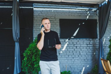 Şirin bir genç adam elinde dizüstü bilgisayarla bir kafede bir terasta duruyor ve telefonla konuşuyor. Çevrimiçi çalış, serbest çalışan. Çevrimiçi uzak yaz işi.