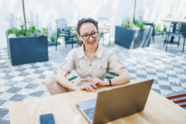 Uzaktan kumanda. Kafede açık terasta oturan rahat bir kadın kahve ve telefonla internette çalışıyor. Serbest çalışan bir kadın, bir yazlık kafede dizüstü bilgisayarın başında otururken uzaktan çalışıyor.