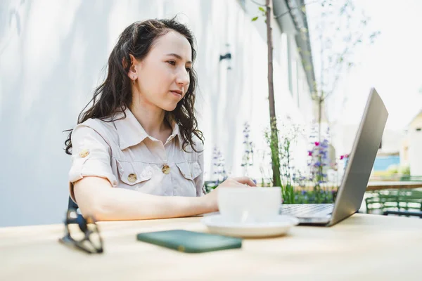 一名女生坐在咖啡馆的夏季阳台上 在笔记本电脑上工作 女自由职业者坐在一家夏季咖啡馆里 在网上远程工作 远程工作 — 图库照片
