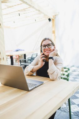 Dışarıdaki kafe masasında dizüstü bilgisayarla oturan gülümseyen kadın, telefonla konuşan ve kahve içen, bardaklı gülümseyen kadın kafede gezmekten ya da internetten ders çalışmaktan zevk alan kadın.