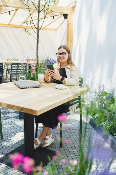 stock image A woman manager sits on an outdoor terrace in a cafe and works online with coffee. Female freelancer works remotely online while sitting in a summer cafe. Remote work