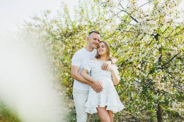 Romantik bir yerde hamilelik bekleyen genç ve güzel bir çift. Çiçek açan bir elma bahçesi. Bahçede yürürken eğlenen mutlu çift. Bir erkek bir kadının elini tutar.