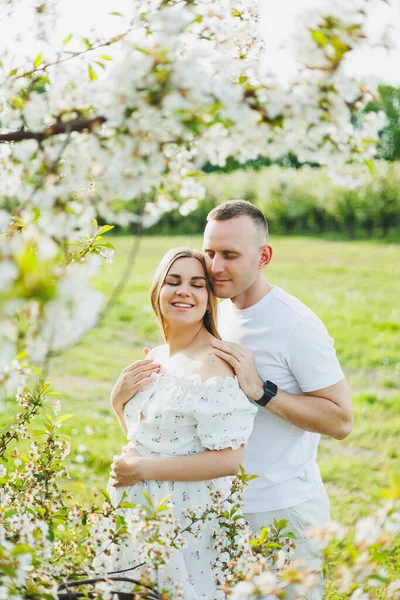 A beautiful young couple expecting a pregnancy in a romantic place, a spring blossoming apple orchard. Happy joyful couple enjoying each other while walking in the garden. A man holds a woman\'s hand