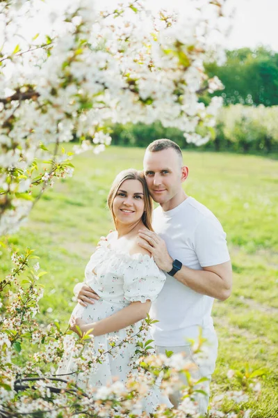 A beautiful young couple expecting a pregnancy in a romantic place, a spring blossoming apple orchard. Happy joyful couple enjoying each other while walking in the garden. A man holds a woman's hand