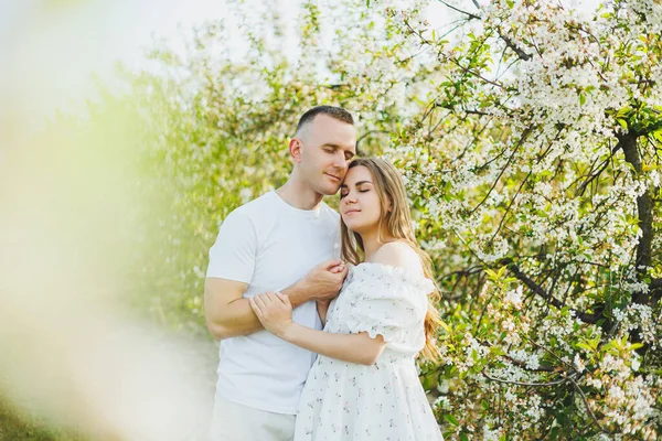 A beautiful young couple expecting a pregnancy in a romantic place, a spring blossoming apple orchard. Happy joyful couple enjoying each other while walking in the garden. A man holds a woman\'s hand