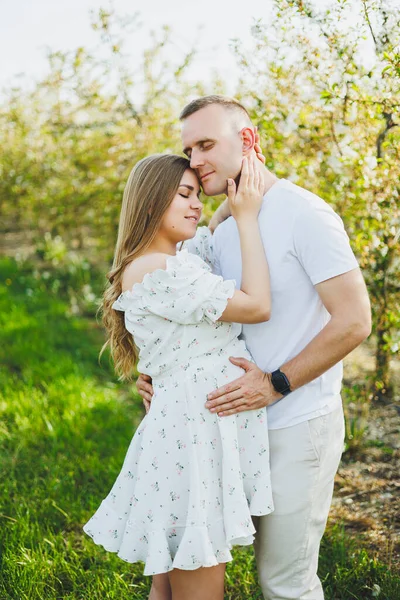 A beautiful young couple expecting a pregnancy in a romantic place, a spring blossoming apple orchard. Happy joyful couple enjoying each other while walking in the garden. A man holds a womans hand