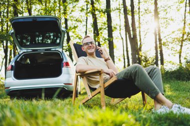 Gözlüklü neşeli bir adam doğada bir sandalyede oturur ve tatilde çalışır, taşınabilir bir dizüstü bilgisayarda çalışır. Serbest çalışma..