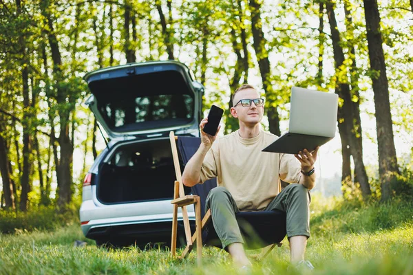 stock image A cheerful man in glasses sits on a chair in nature and works on vacation, work online on a portable laptop. Freelance work.