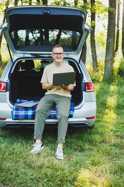 stock image Young smiling man in glasses freelancer works on a laptop while sitting in the trunk of a car. Remote work in nature.