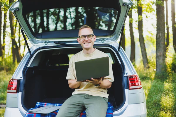 Young smiling man in glasses freelancer works on a laptop while sitting in the trunk of a car. Remote work in nature.