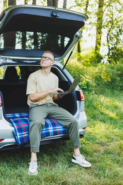 Young smiling man in glasses freelancer works on a laptop while sitting in the trunk of a car. Remote work in nature.