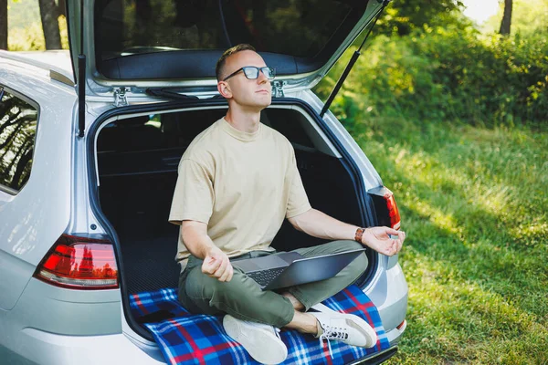 stock image Young smiling man in glasses freelancer works on a laptop while sitting in the trunk of a car. Remote work in nature.