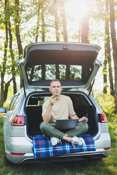 stock image Young smiling man in glasses freelancer works on a laptop while sitting in the trunk of a car. Remote work in nature.