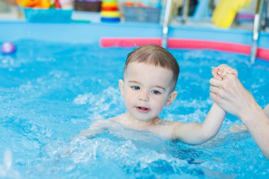 2 yaşındaki bir çocuk bir koçla havuzda yüzmeyi öğreniyor. Çocuklar için yüzme dersleri. Çocuklar için yüzme okulu. Çocuklar için eğitici yüzme dersleri