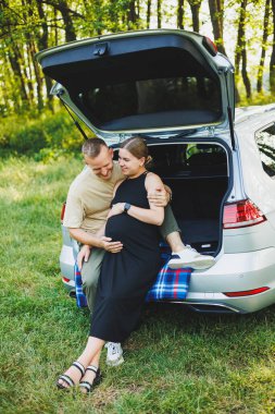 Çocuk bekleyen mutlu, evli, genç bir çift doğada bir arabanın bagajında oturuyor. Bir erkek ve bir kadın, yeşil bir çimenlikte otururken hamile bir göbeğe sarılıyorlar.